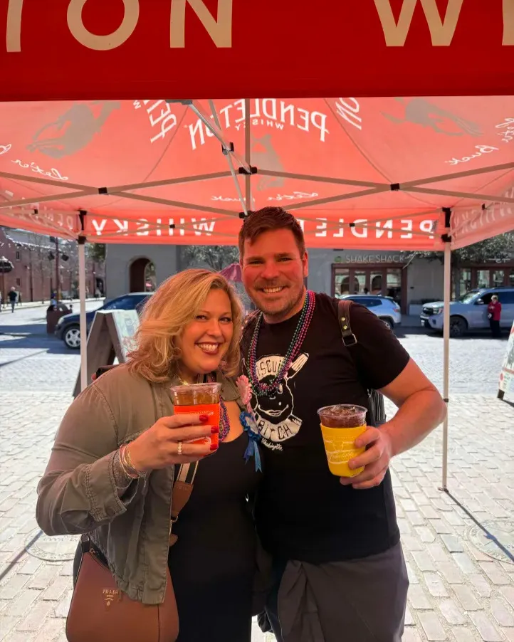 A smiling couple holding colorful drinks under a red whiskey tent on a Stockyards brick street