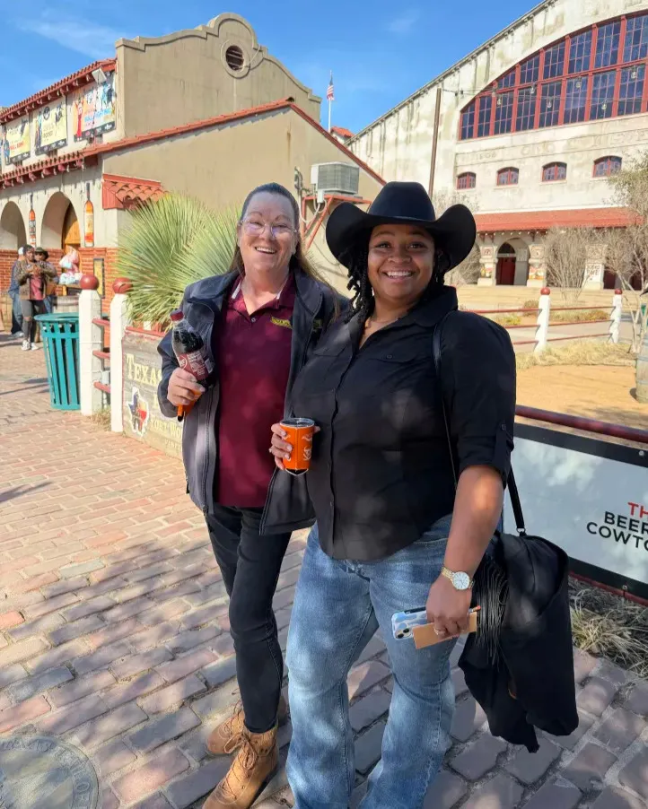Two smiling guests, one in a black cowboy hat, holding drinks in front of Stockyards buildings